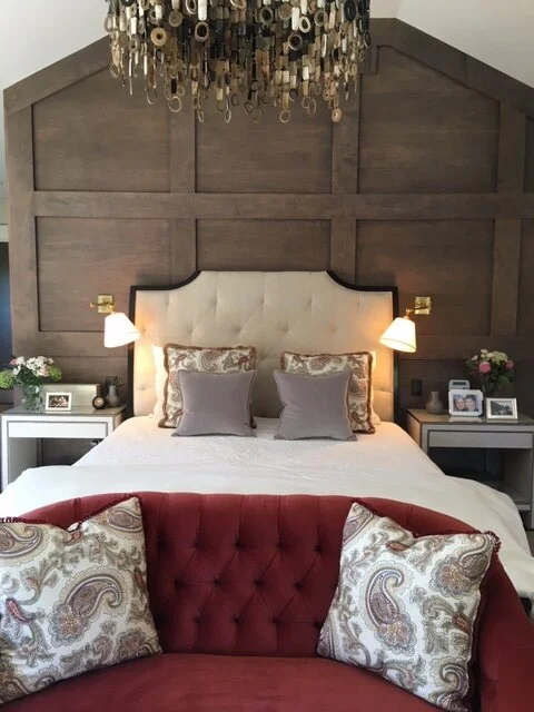 The deep stained oak paneled wall in this room creates the perfect backdrop for the upholstered headboard and transitional tables. The rich color palette of Lavender and deep red only build on the luxury with an eclectic touch with the horn chandelier completing the look.